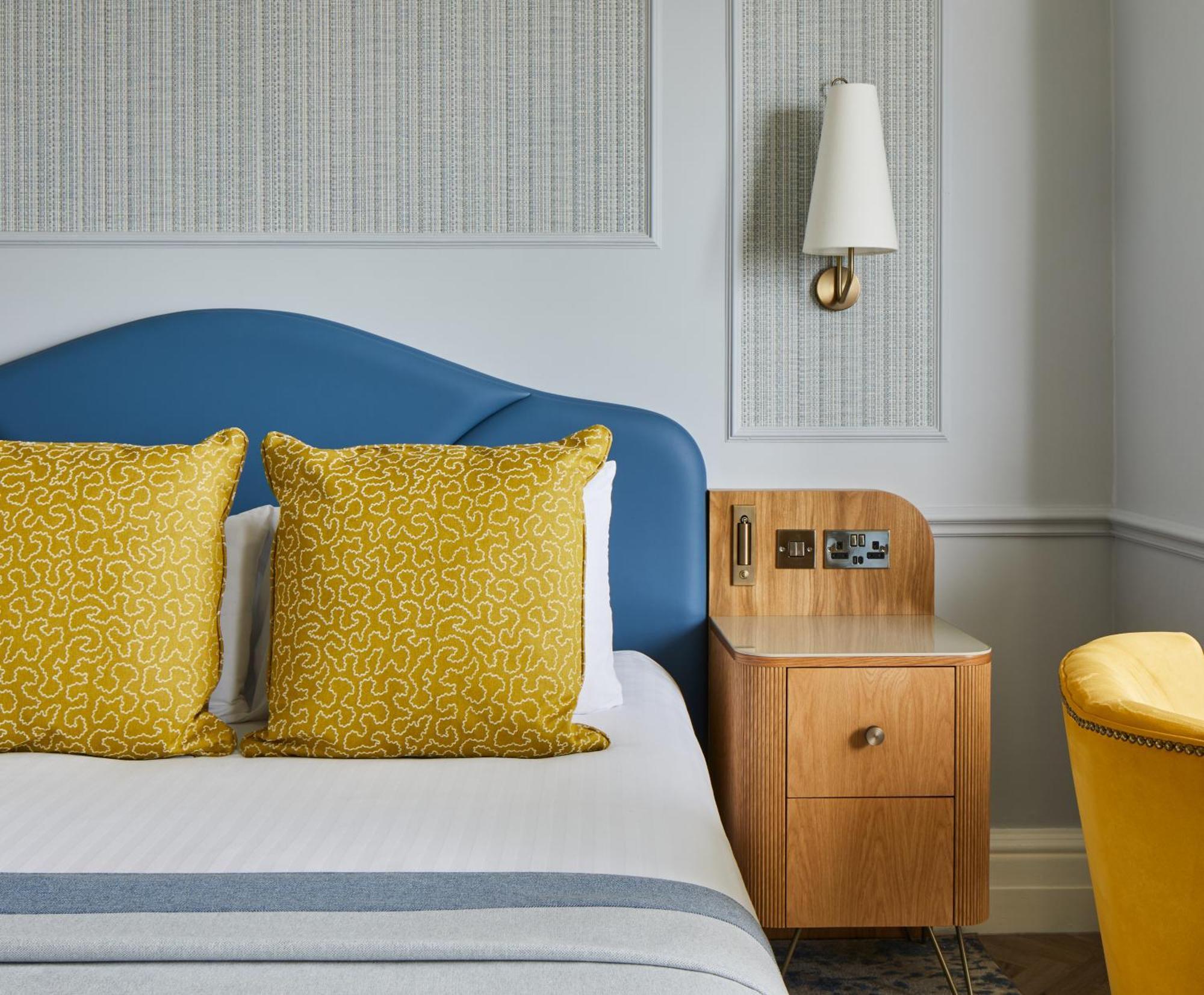 The Grand Brighton Hotell Exteriör bild The photo shows a bedroom with a modern and stylish design. There is a bed with a curved blue headboard and two yellow decorative pillows on it. Next to the bed, there is a wooden nightstand featuring an electrical outlet and a drawer. On the wall ab