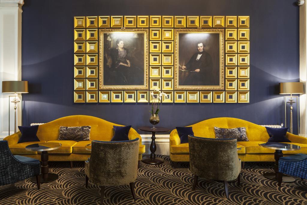 The Grand Brighton Hotell Exteriör bild The photo depicts an elegant interior space. Two large, ornate portraits are prominently displayed in a decorative gold frame on a dark blue wall. Below the portraits, there are two yellow sofas complemented by plush armchairs in a neutral tone. The 